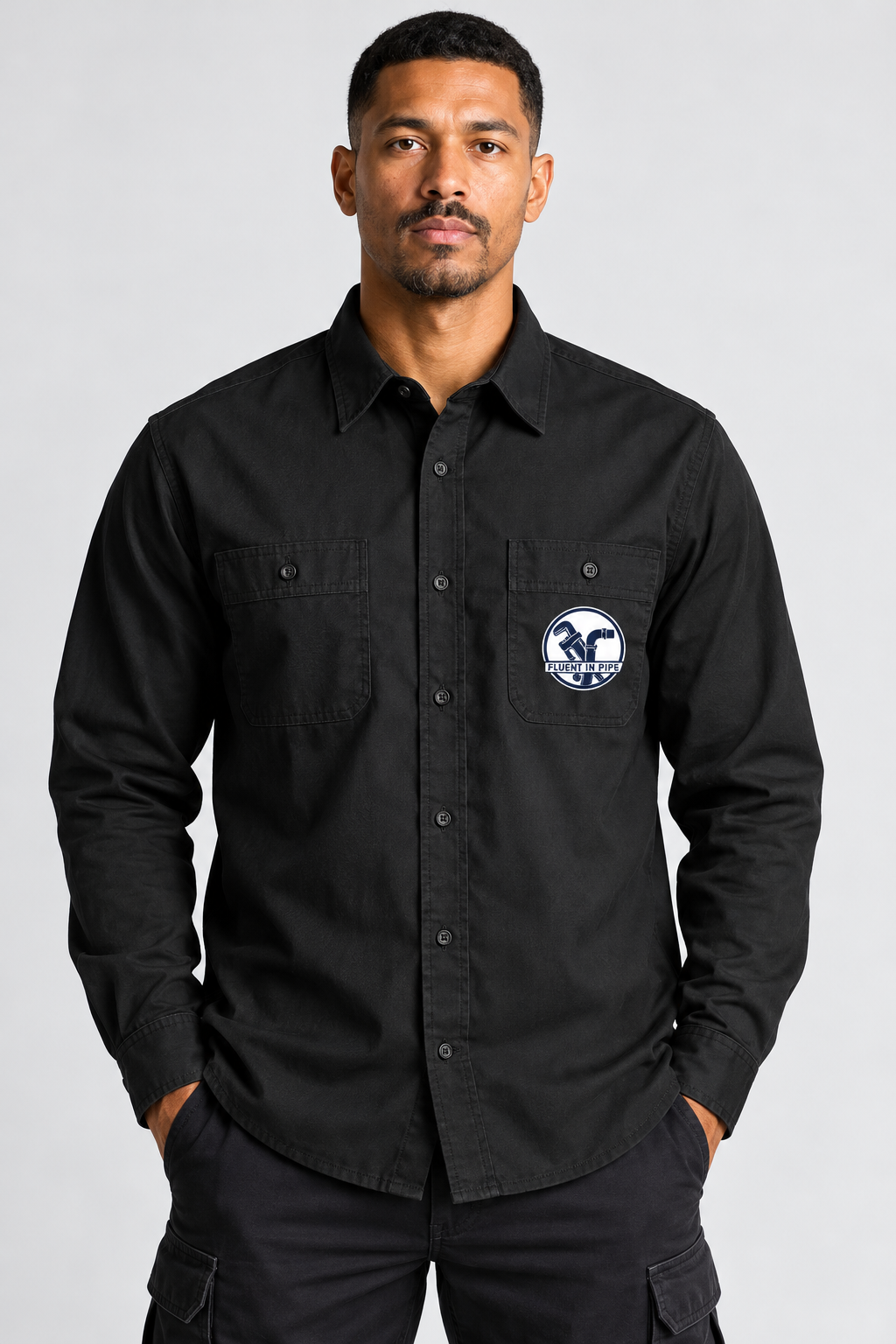A man wearing a black long-sleeve button-up shirt with a circular blue and white logo patch on the left chest pocket, posed against a light gray studio background.